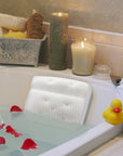 A relaxing and comfortable bathtub setup with a grace & stella bath pillow, rose petals, lit candles, and a rubber duck.