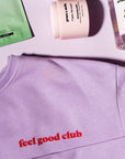 Lilac crewneck sweater from grace & stella's "Feel Good Club" line, accompanied by skincare products on a pink surface.
