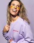 Woman with a smile wearing gold under-eye patches and an oversized grace & stella lilac crew neck sweater with the text "Feel Good Club".