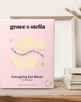 Pink package of grace & stella free sample set of eye masks (6 pairs) for dark circles placed on a wooden shelf next to a white vase and framed artwork.