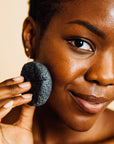 A woman holding a konjac facial cleansing sponge from grace & stella co. near her cheek.