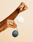 Two hands holding a white and black grace & stella co. Konjac Facial Cleansing Sponge against a pale background.