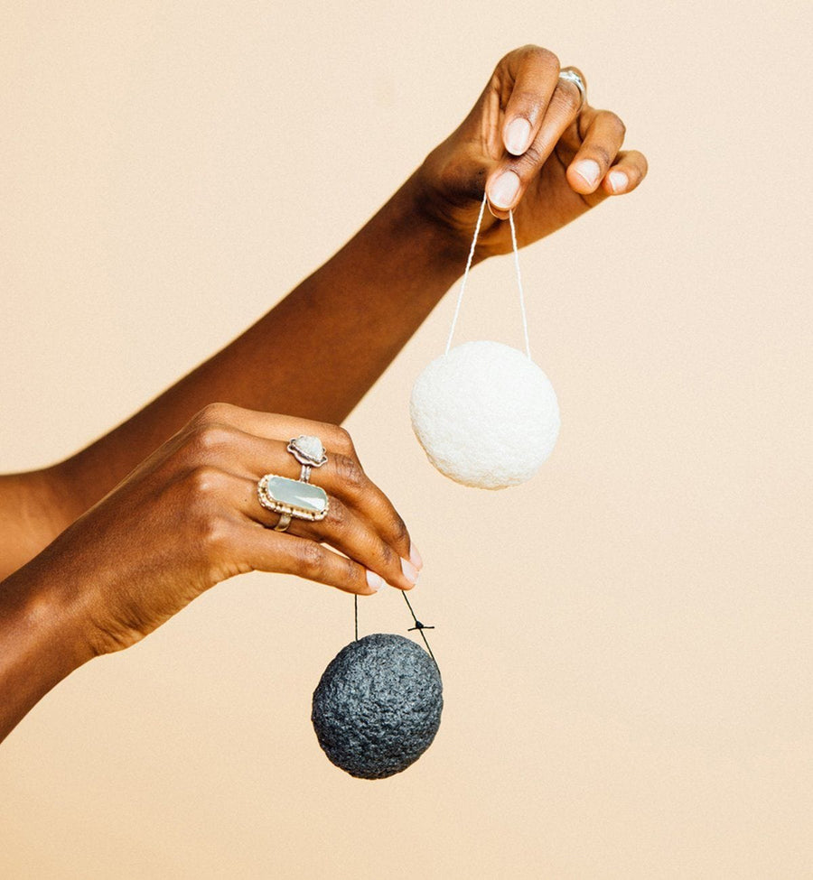 Two hands holding a white and black grace & stella co. Konjac Facial Cleansing Sponge against a pale background.