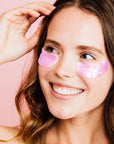 A smiling woman with Grace & Stella La Vie En Rose Set under-eye patches on a fresh pink background.