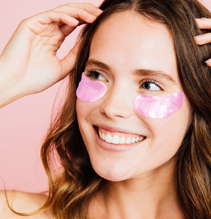 A smiling woman with Grace & Stella La Vie En Rose Set under-eye patches on a fresh pink background.