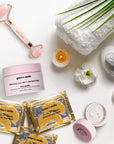 Flat lay of skincare products with a jade roller, towels, stones, candle, eye cream from grace & stella's energizing set and a plant on a white background.