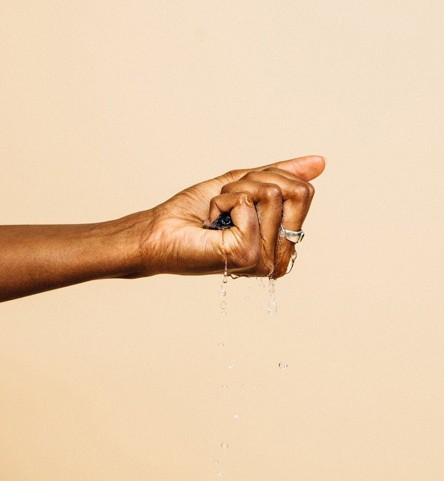 A person's hand squeezing a Grace & Stella Co. Konjac Facial Cleansing Sponge, with water droplets visible against a plain background.