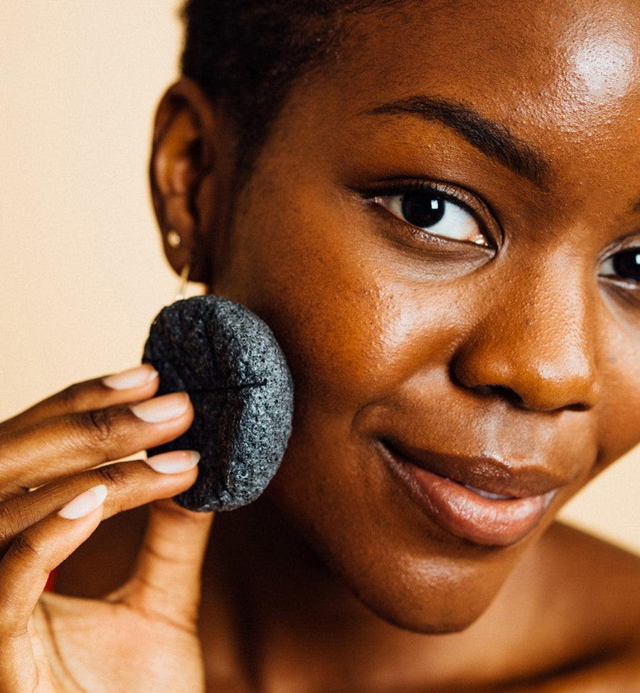 A woman holding a konjac facial cleansing sponge from grace & stella co. near her cheek.