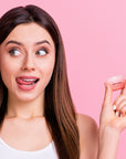 A young woman is holding up a Grace & Stella La Vie En Rose Set lip balm on a pink background for touch-ups.